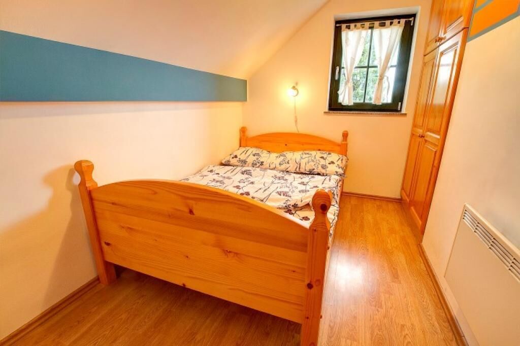 A small bedroom with a wooden bed, a window with curtains, and a wall-mounted light.