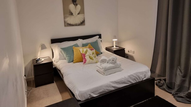 A neatly made bed with white linens and a few pillows, a bedside lamp, and a small table with a lamp. - Spacious 2 Bedroom Duplex (Reading, Berkshire)