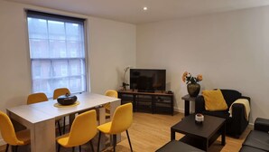 A modern living room with a dining area, a flat-screen TV, and a sofa with a yellow throw pillow. - Spacious 2 Bedroom Duplex (Reading, Berkshire)