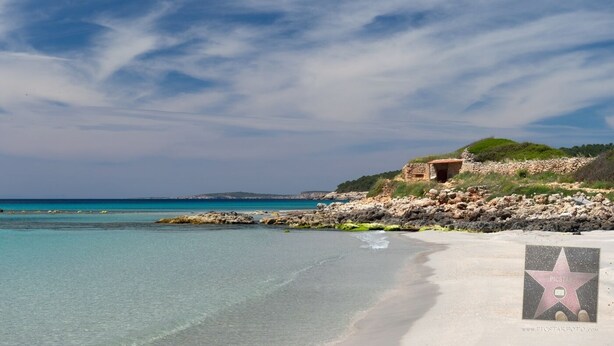 A beach with clear turquoise waters, a rocky shoreline, and a small building on a hill.