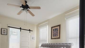 Une chambre avec un lit, un ventilateur de plafond, une fenêtre avec des stores, un tableau encadré au mur et une porte avec un porte-serviettes.