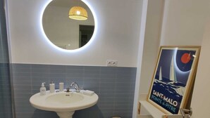 Un baño con un espejo redondo, un lavabo pedestal blanco y un cartel enmarcado en la pared.