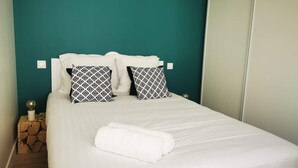Una cama bien tendida con ropa de cama blanca y almohadas estampadas, una pared color turquesa y una mesita de noche de madera con una lámpara.