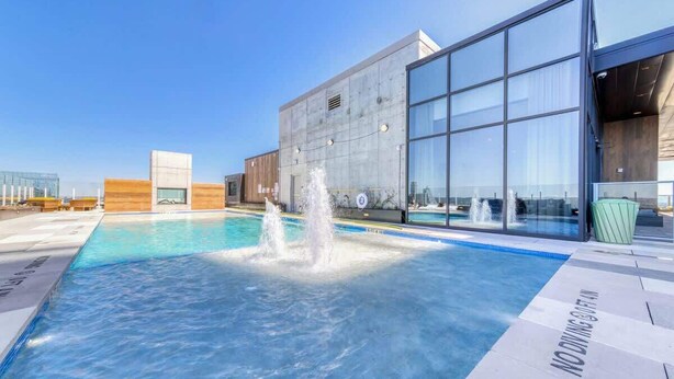 A swimming pool with a fountain, a modern building with large windows, and a clear sky. - Frisco - Rooftop pool & gym on Rainey St (Austin)