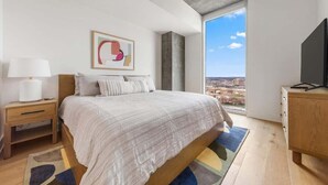 A modern bedroom with a large bed, a nightstand, a dresser, a wall-mounted TV, and a city view through a large window. - Frisco - Rooftop pool & gym on Rainey St (Austin)