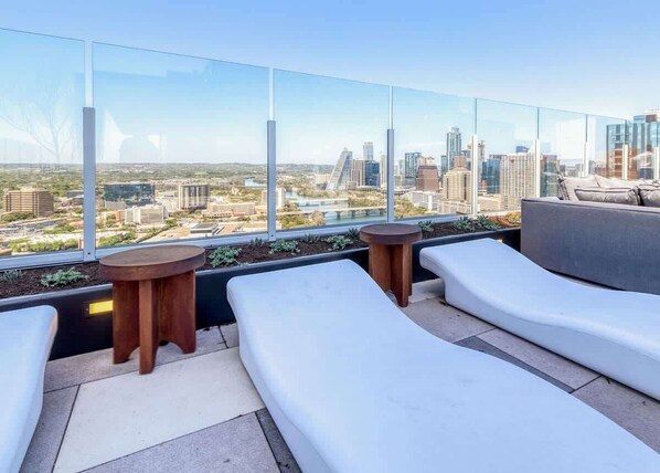 A rooftop lounge with white cushioned seating, a round wooden table, and a glass railing overlooking a cityscape.