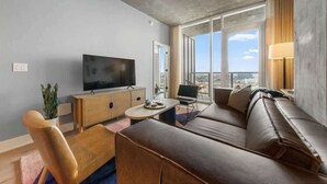 A modern hotel room with a large window, a flat-screen TV, a sofa, a chair, and a small table. - Frisco - Rooftop pool & gym on Rainey St (Austin)