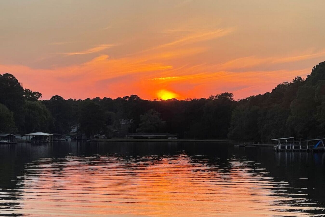 A sunset over a lake with a forested shoreline and buildings in the background.