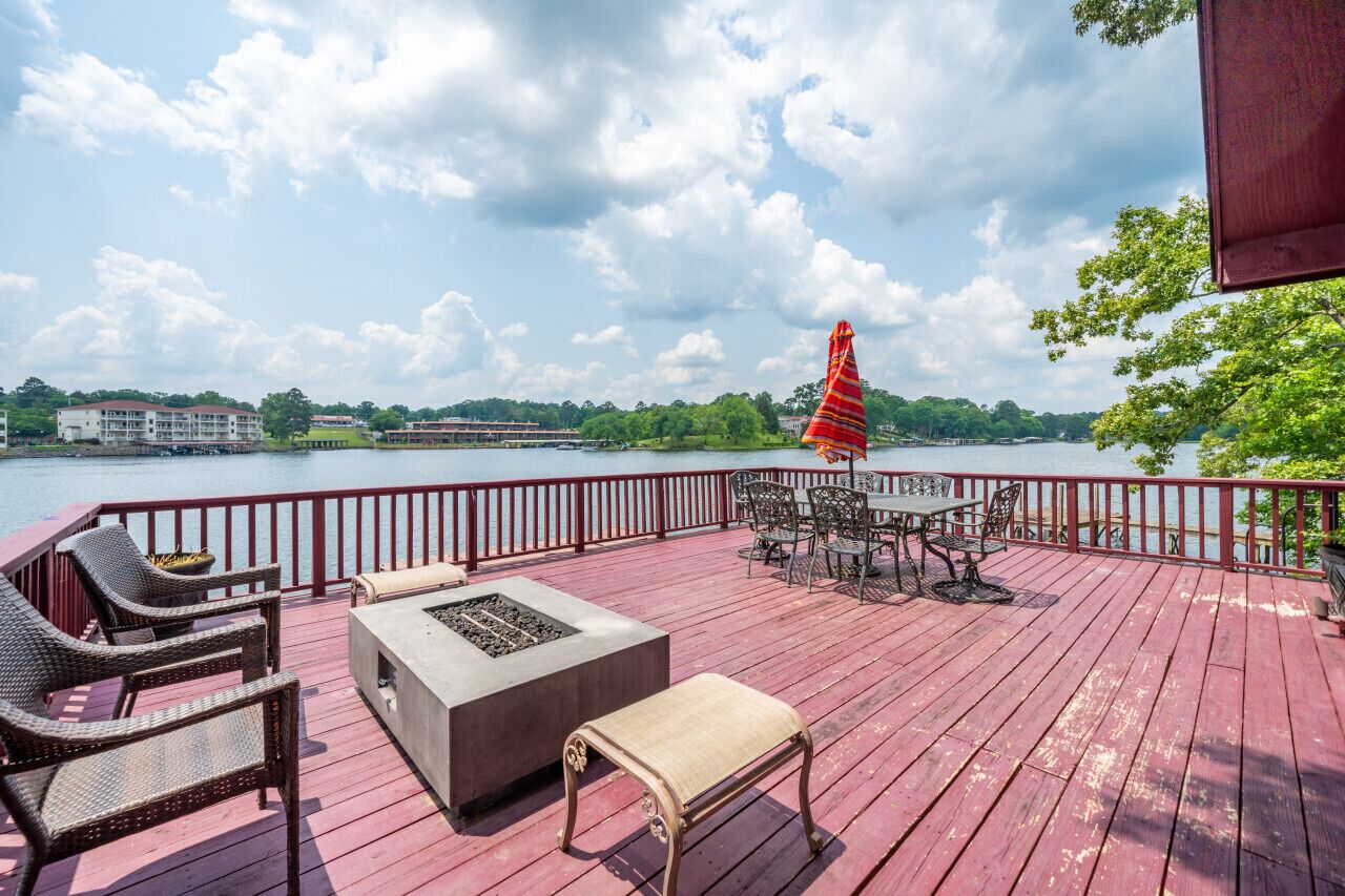 Une terrasse en bois avec des meubles d’extérieur, un foyer et une vue sur un lac et des bâtiments en arrière-plan.