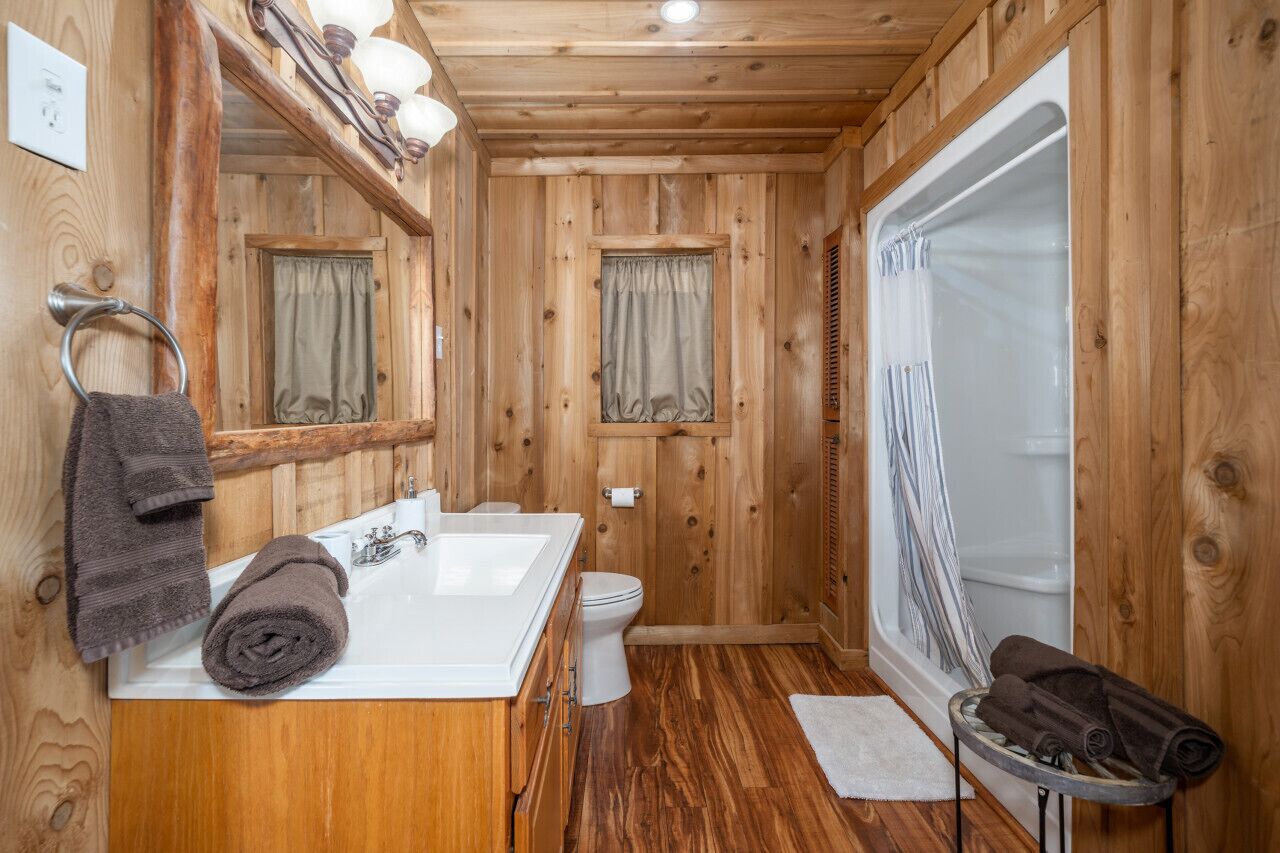 Een houten badkamer van een chalet, met een witte wastafel, twee ramen met gordijnen, een toilet en een douchehoek.