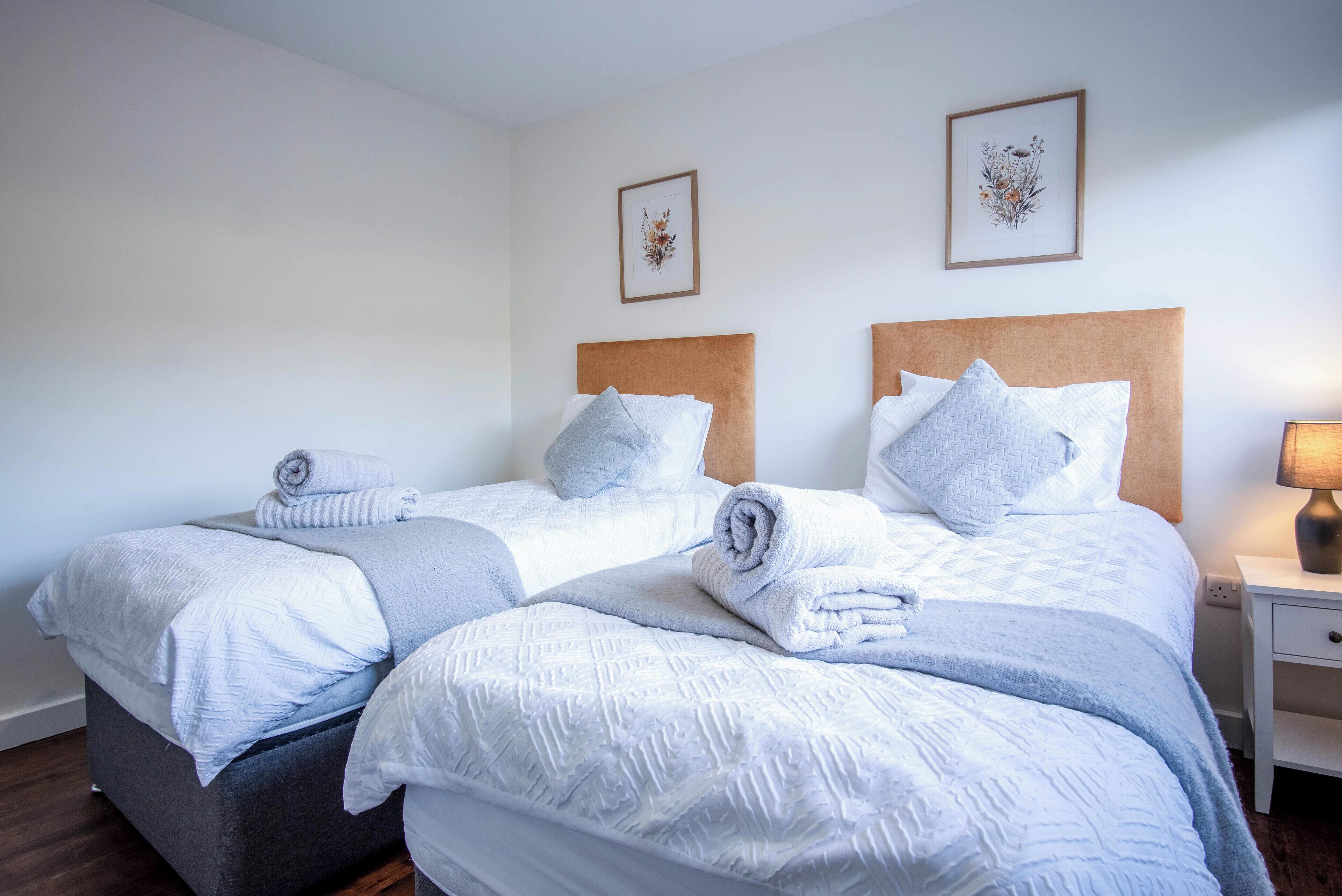 Hotellihuoneessa on kaksi sänkyä, joissa on molemmissa pääty sekä tyynyt ja rullalle taitetut pyyhkeet. Seinällä on kehystettyjä kuvia ja oikealla puolella yöpöydällä on lamppu.