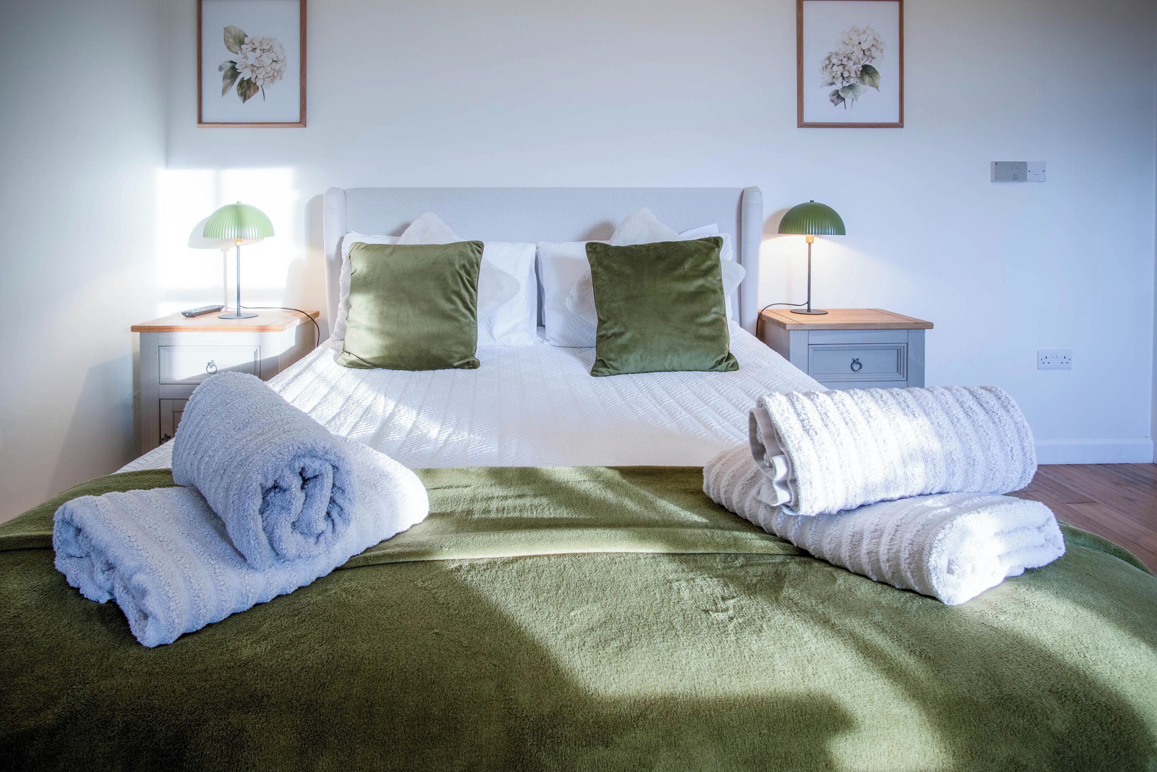 A neatly made bed with green and white bedding, two bedside lamps, and a framed floral artwork on the wall.