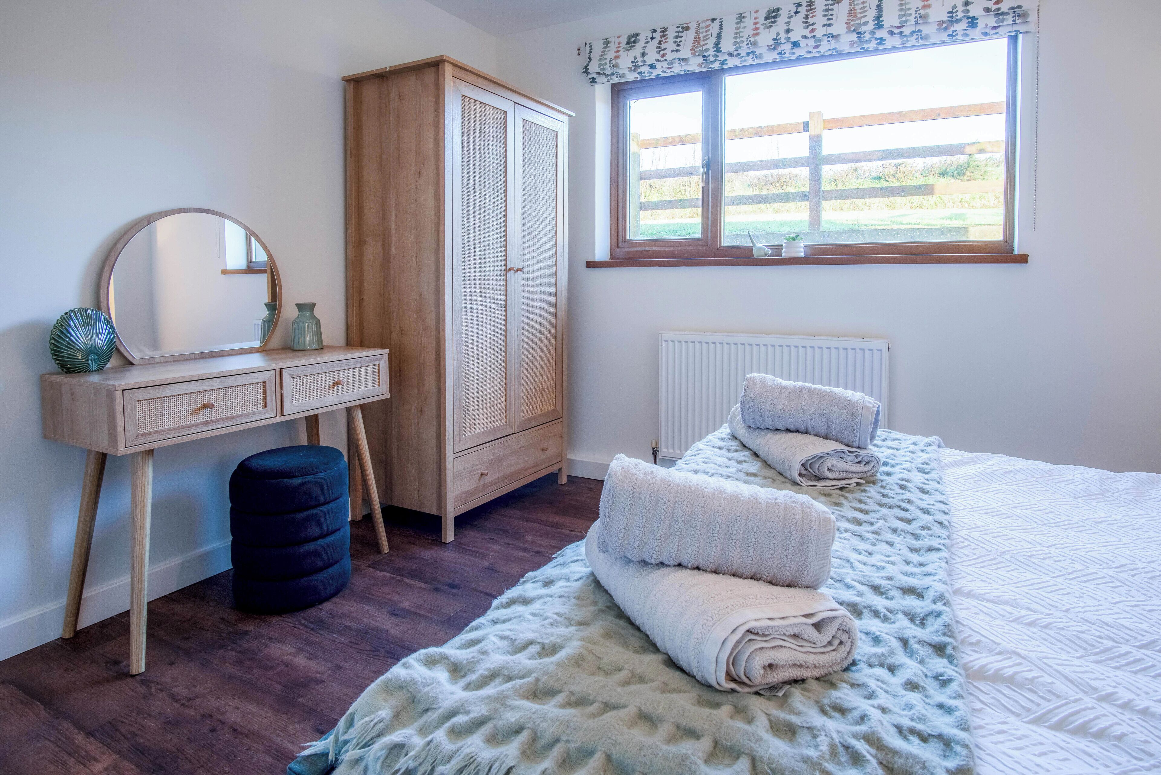 Een slaapkamer met een houten garderobe, een ronde spiegel, een nachtkastje, een radiator en een raam met uitzicht op de omgeving.