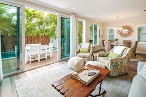A spacious living room with a large sliding glass door leading to a patio, furnished with a white bench, wicker chairs, and a coffee table.