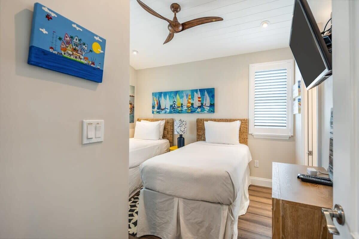 A hotel room with two beds, a ceiling fan, a painting of sailboats, and a television on a wooden cabinet.