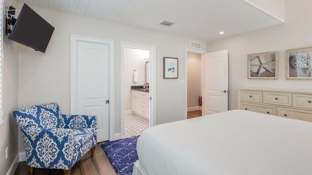 Une chambre à coucher avec un lit, une chaise, un téléviseur et une salle de bain visible à travers une porte ouverte.