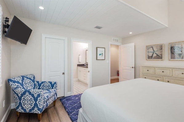 Une chambre à coucher avec un lit, une chaise, un téléviseur et une salle de bain visible à travers une porte ouverte.