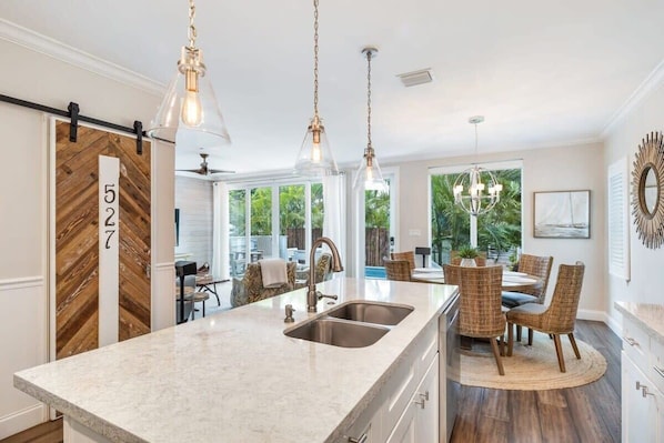 A kitchen with a large island, stainless steel sink, pendant lighting, and a dining area with wicker chairs.