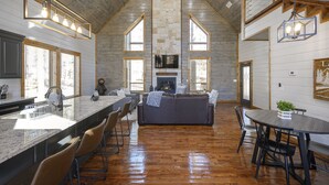 A spacious kitchen with a long island, a fireplace, and a dining area with a round table and chairs. - Sunrise Retreat - Hot Tub, 2 Story Deck, Pet Friendly, Free Firewood (Broken Bow)
