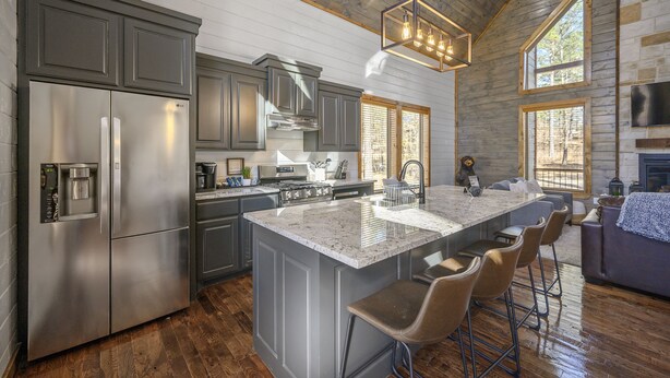 A modern kitchen with stainless steel appliances, a large island with bar stools, and a cozy seating area with a fireplace. - Sunrise Retreat - Hot Tub, 2 Story Deck, Pet Friendly, Free Firewood (Broken Bow)