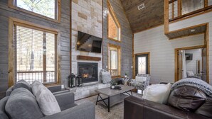 A cozy living room with a fireplace, large windows, and a stone wall. - Sunrise Retreat - Hot Tub, 2 Story Deck, Pet Friendly, Free Firewood (Broken Bow)