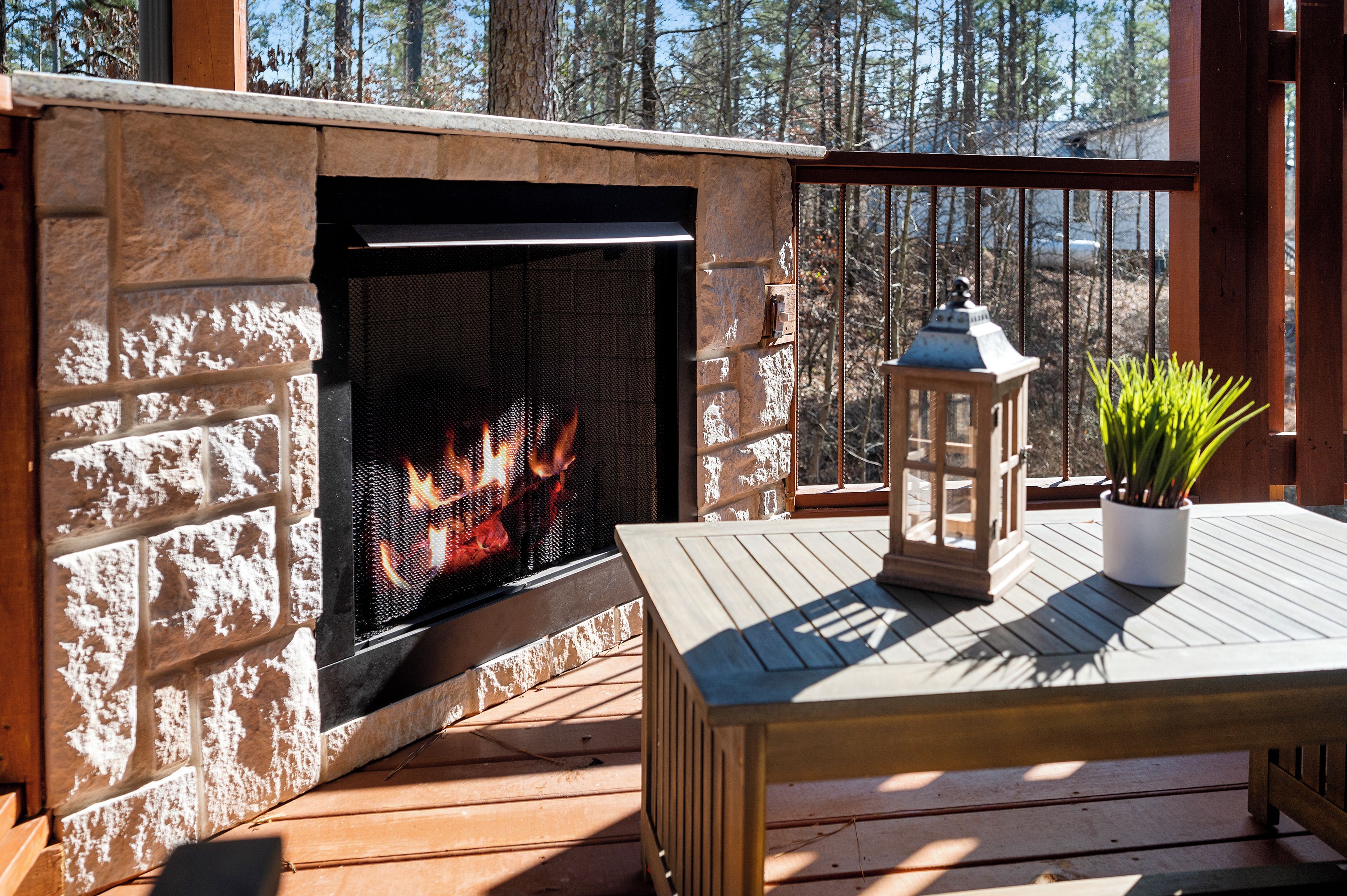 A stone fireplace with a fire, a wooden table, a lantern, and a potted plant on a deck.