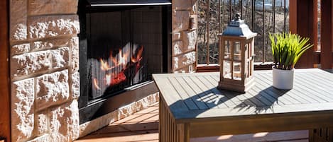 A stone fireplace with a fire, a wooden table, a lantern, and a potted plant on a deck.