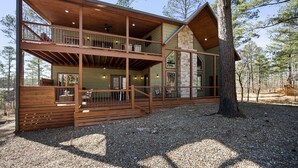 A two-story log cabin with a balcony, large windows, and a stone fireplace. - Sunrise Retreat - Hot Tub, 2 Story Deck, Pet Friendly, Free Firewood (Broken Bow)
