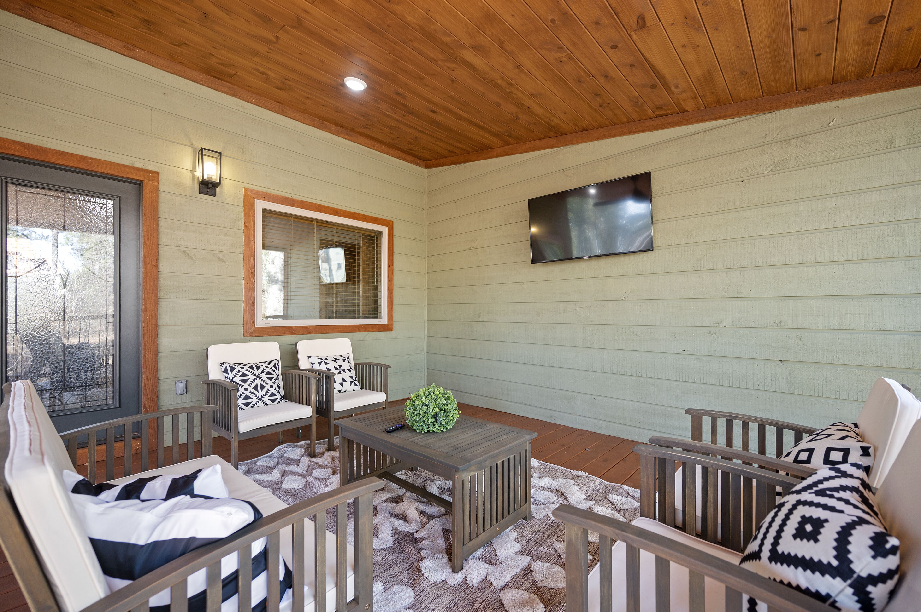 A cozy outdoor seating area with a wooden ceiling, a flat-screen TV mounted on the wall, and a comfortable seating arrangement including a white chair and a bench.