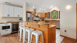 A modern kitchen with white cabinets, a granite countertop, and a central island with bar stools. - Z's By The Sea: Private Pool | Hot Tub | Rec Room (Kitty Hawk)