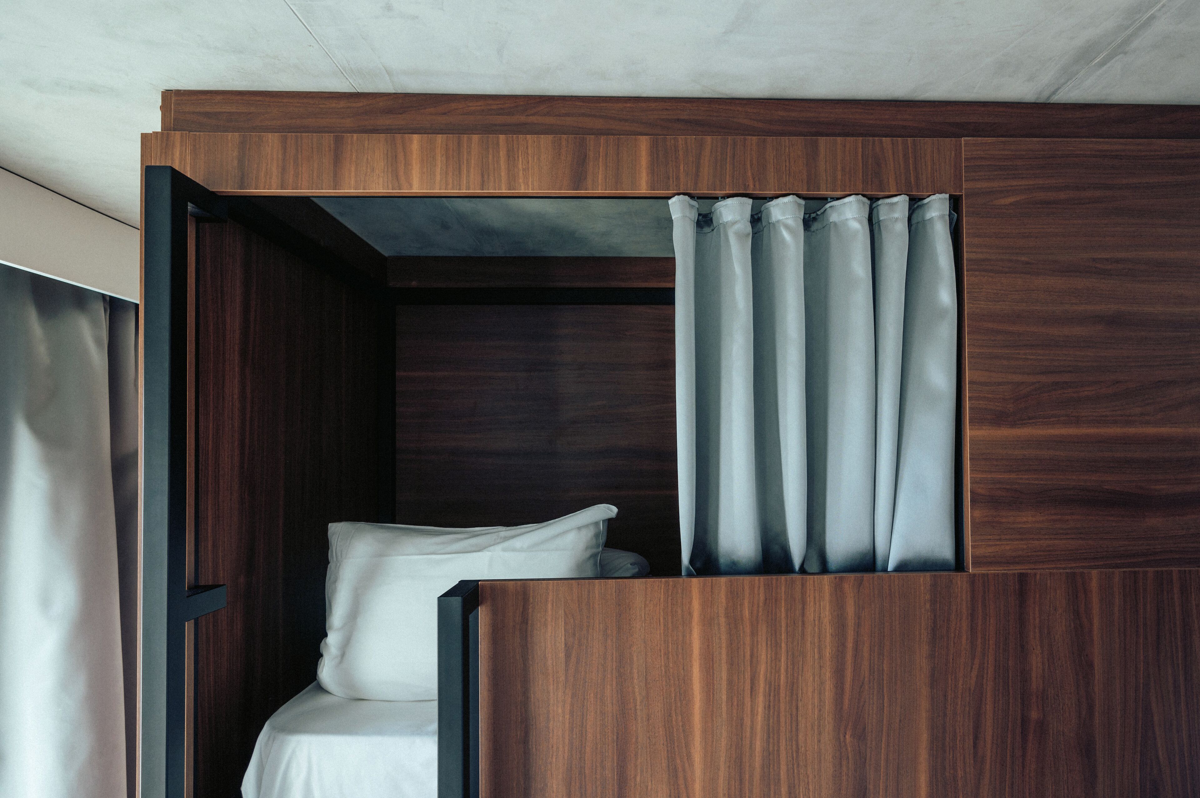 Quarto moderno e compacto com cabeceira de madeira, uma cama de solteiro e uma cortina parcialmente fechada.