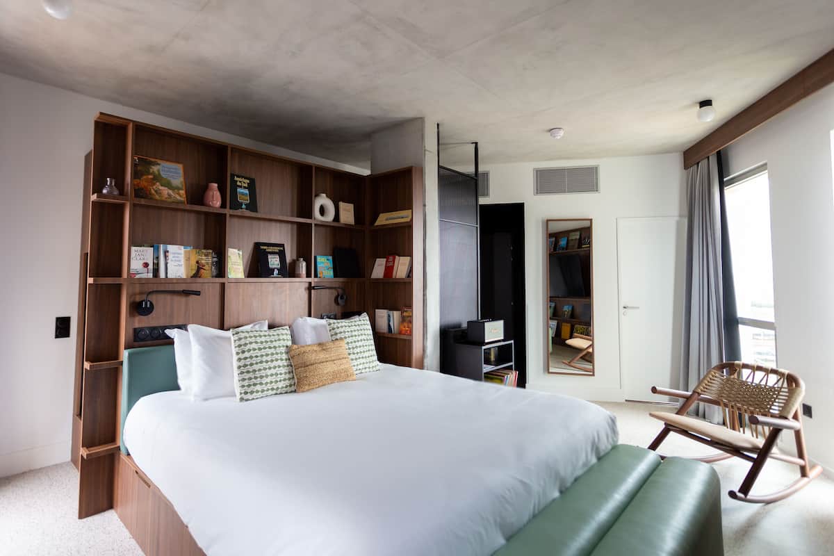 Une chambre moderne avec un grand lit, des étagères intégrées où sont rangés des livres et des objets décoratifs, un fauteuil à bascule et une grande fenêtre avec des rideaux.