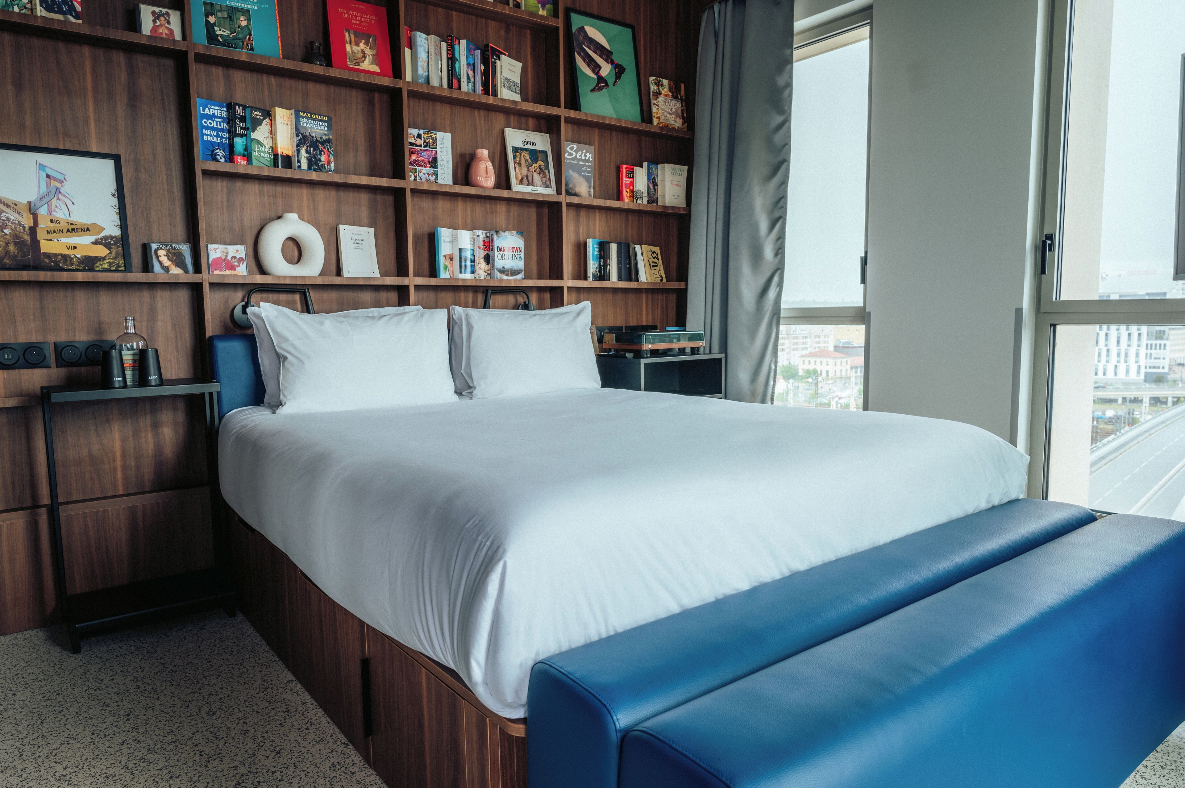 A hotel room with a large bed, a blue bench, wooden shelves with books and decor, and a city view through the window.