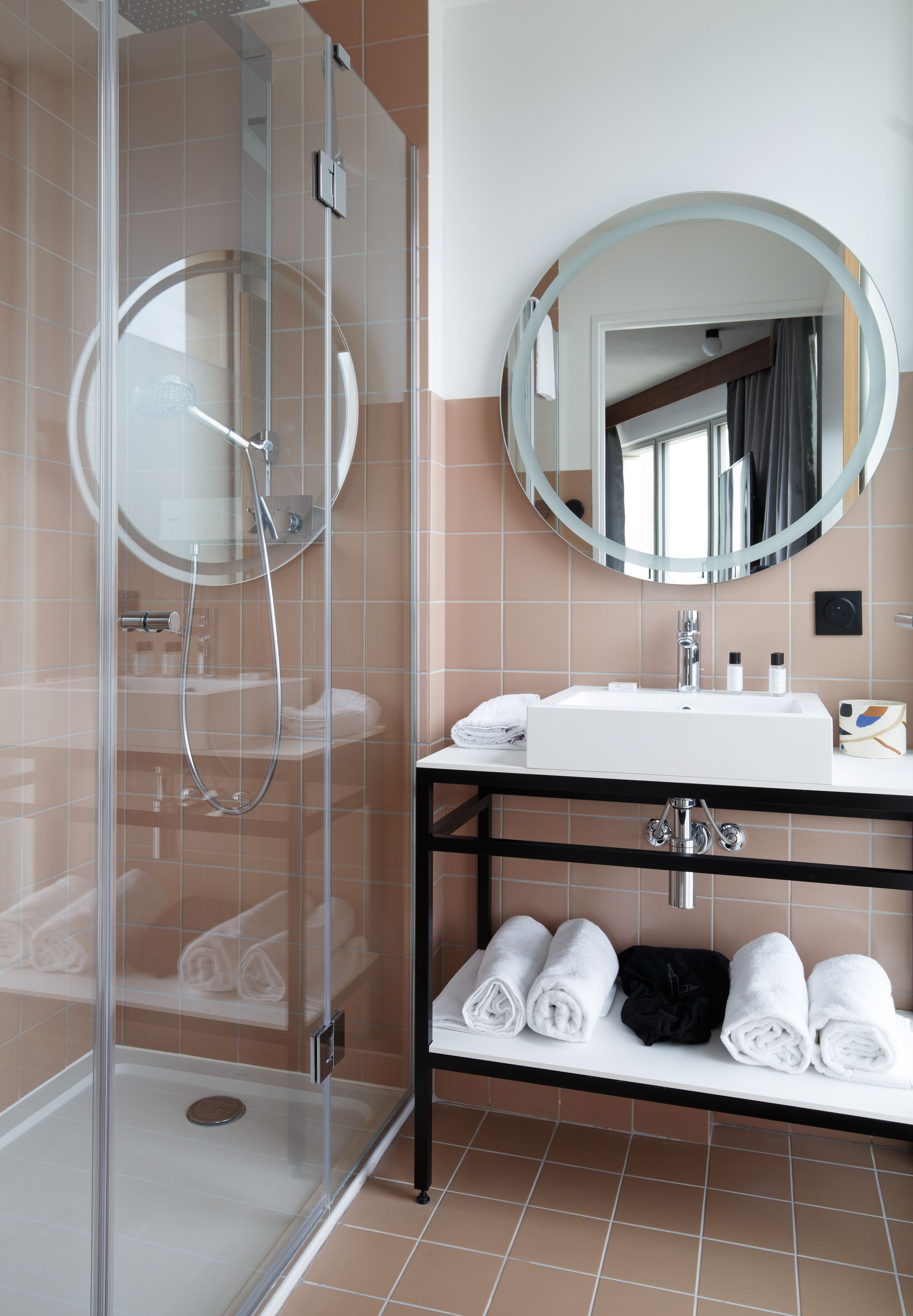 Un baño moderno con ducha de vidrio, un lavamanos blanco, un espejo redondo y toallas dobladas con cuidado en un estante negro.