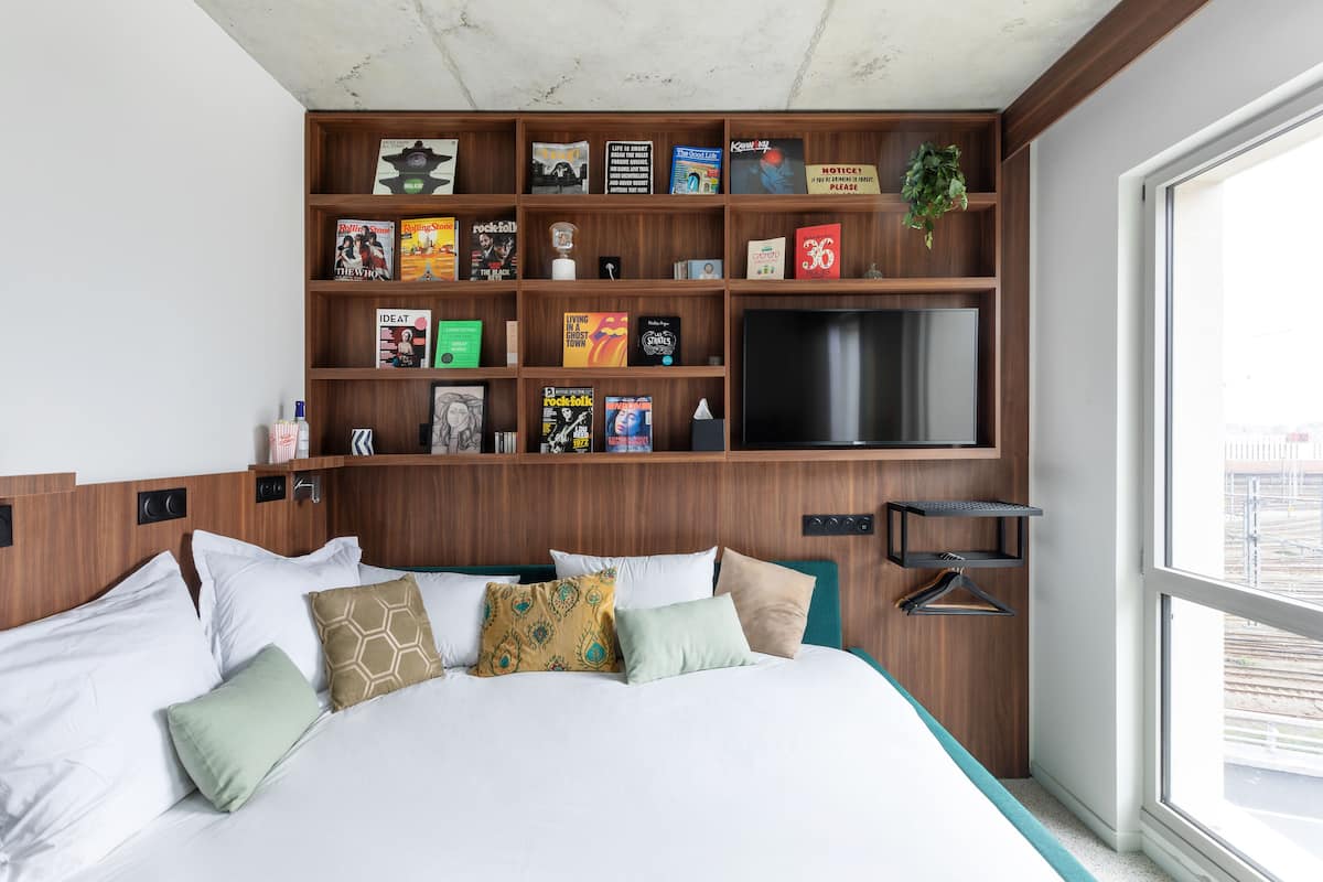 Une chambre d’hôtel avec un grand lit, une bibliothèque en bois remplie de divers ouvrages, un téléviseur à écran plat et une vue sur la ville par une grande fenêtre.
