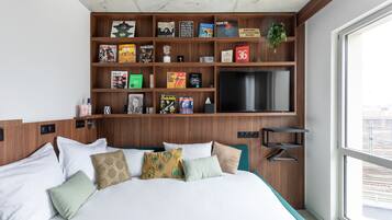 Una habitación de hotel con una cama grande, una estantería de madera con varios libros, un televisor de pantalla plana y vistas a la ciudad a través de un ventanal amplio.