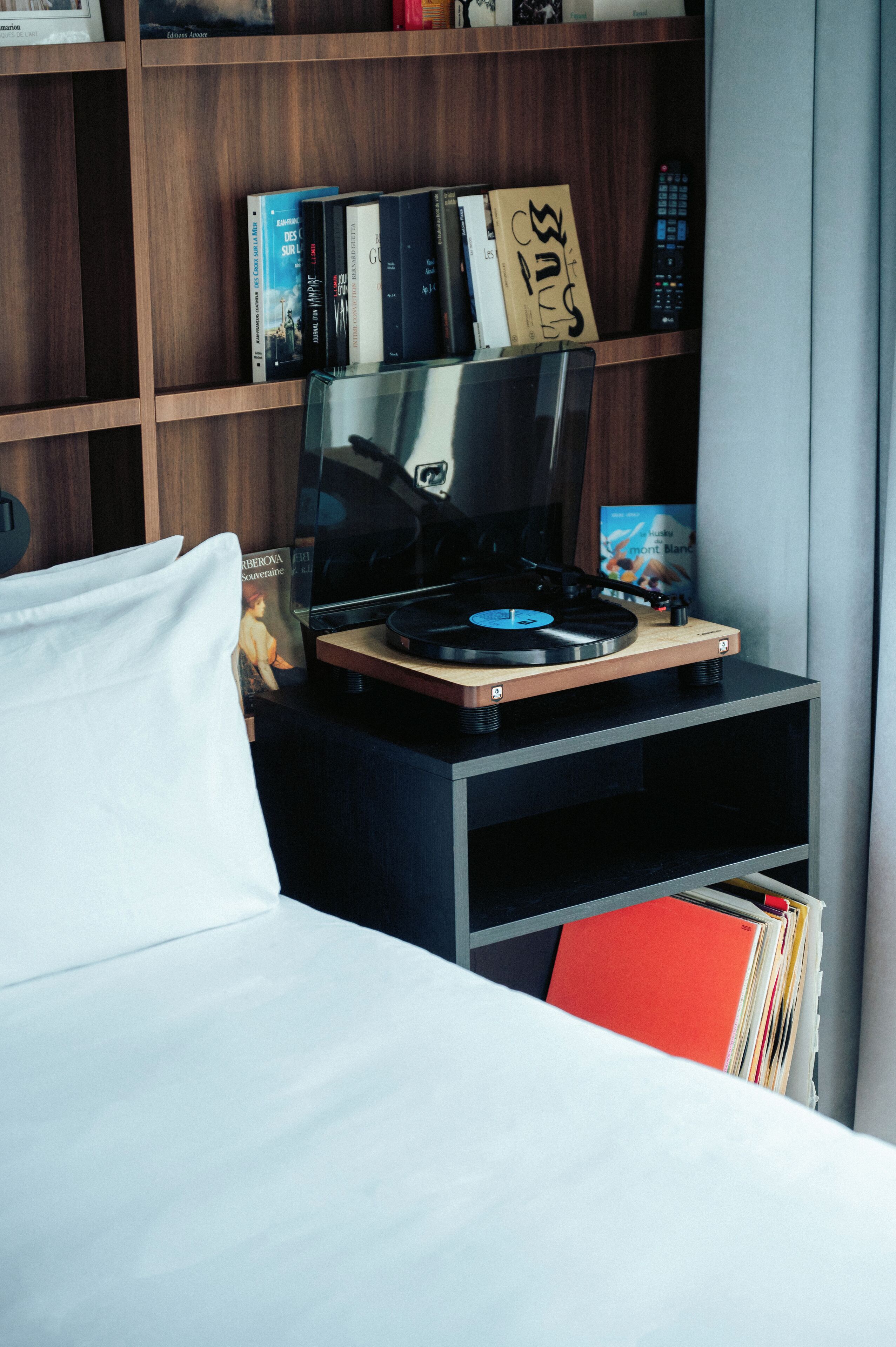 Une chambre moderne avec un lit, une petite table en bois sur laquelle se trouve une platine, une bibliothèque remplie de livres et une télécommande, et un dossier rouge posé sur le sol.