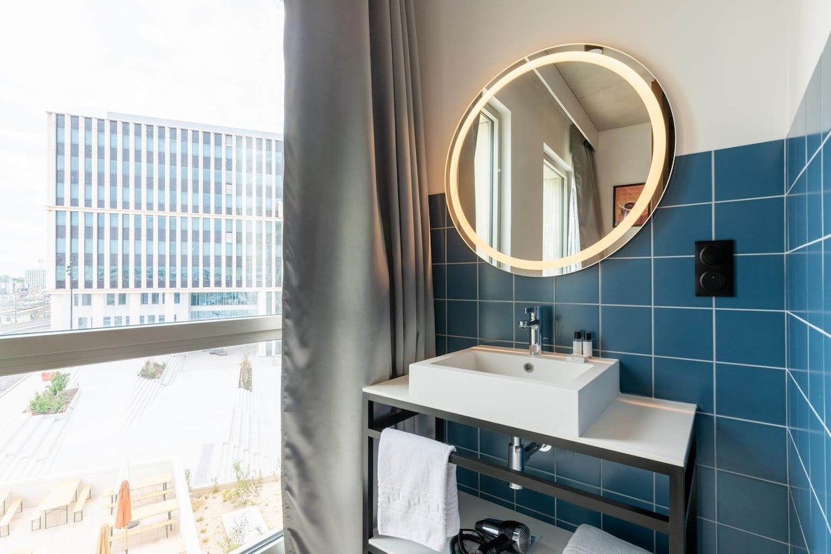 Un baño moderno con un lavabo blanco, un espejo redondo y vistas a un paisaje urbano a través de un ventanal grande.