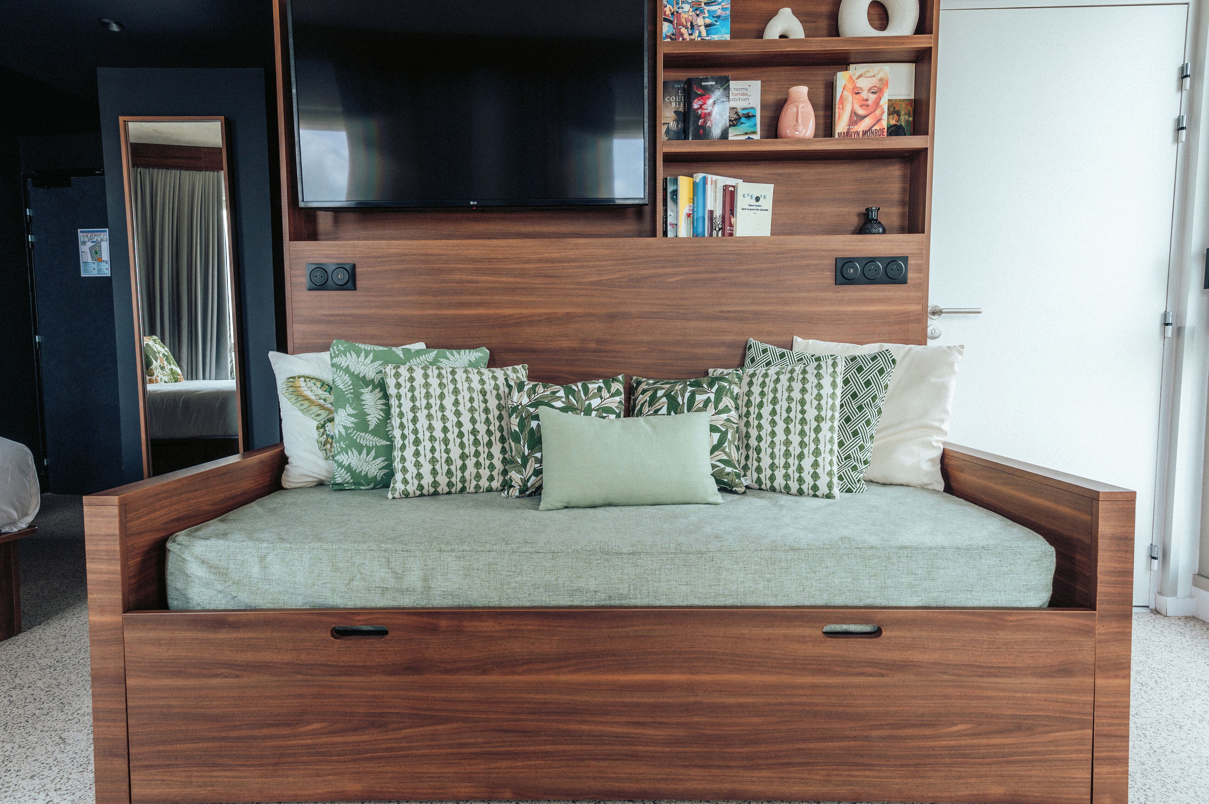 A modern bedroom with a wooden headboard, a flat-screen TV, a bookshelf, and a large window.