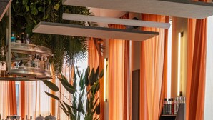 A modern dining area with a bar, wooden tables, and chairs, surrounded by orange curtains and indoor plants. - JOST Hôtel Bordeaux Centre Gare Saint Jean (Bordeaux)