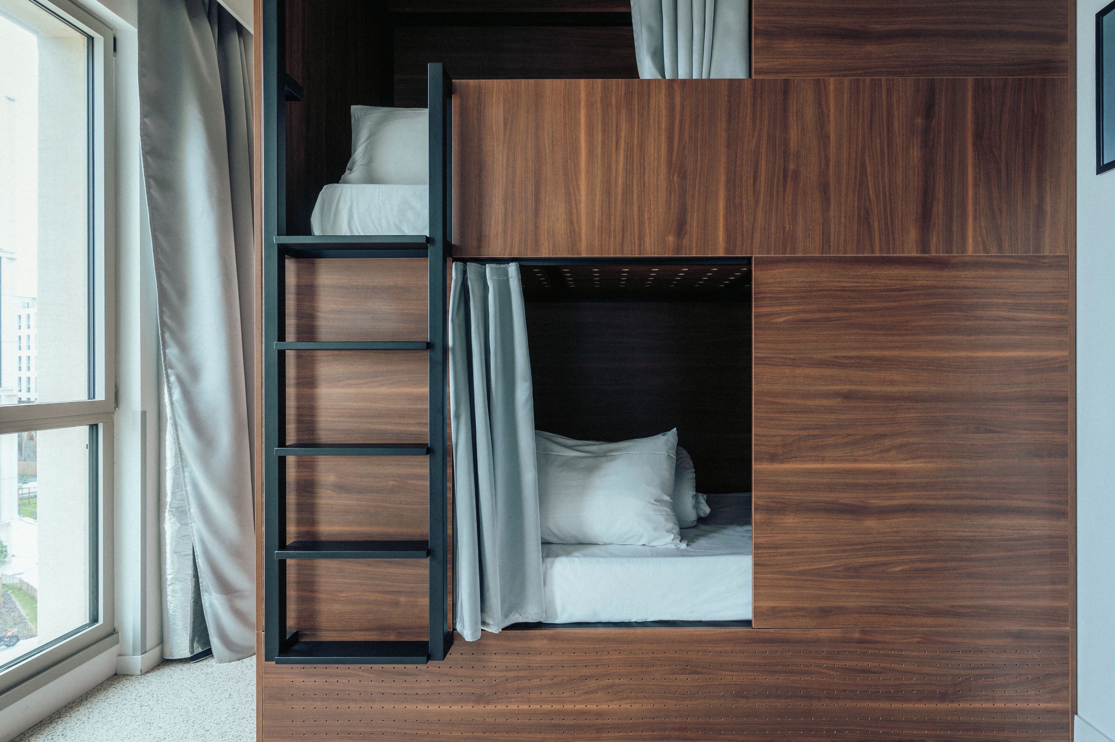 A bunk bed with a wooden frame and a ladder, a window with curtains, and a city view.