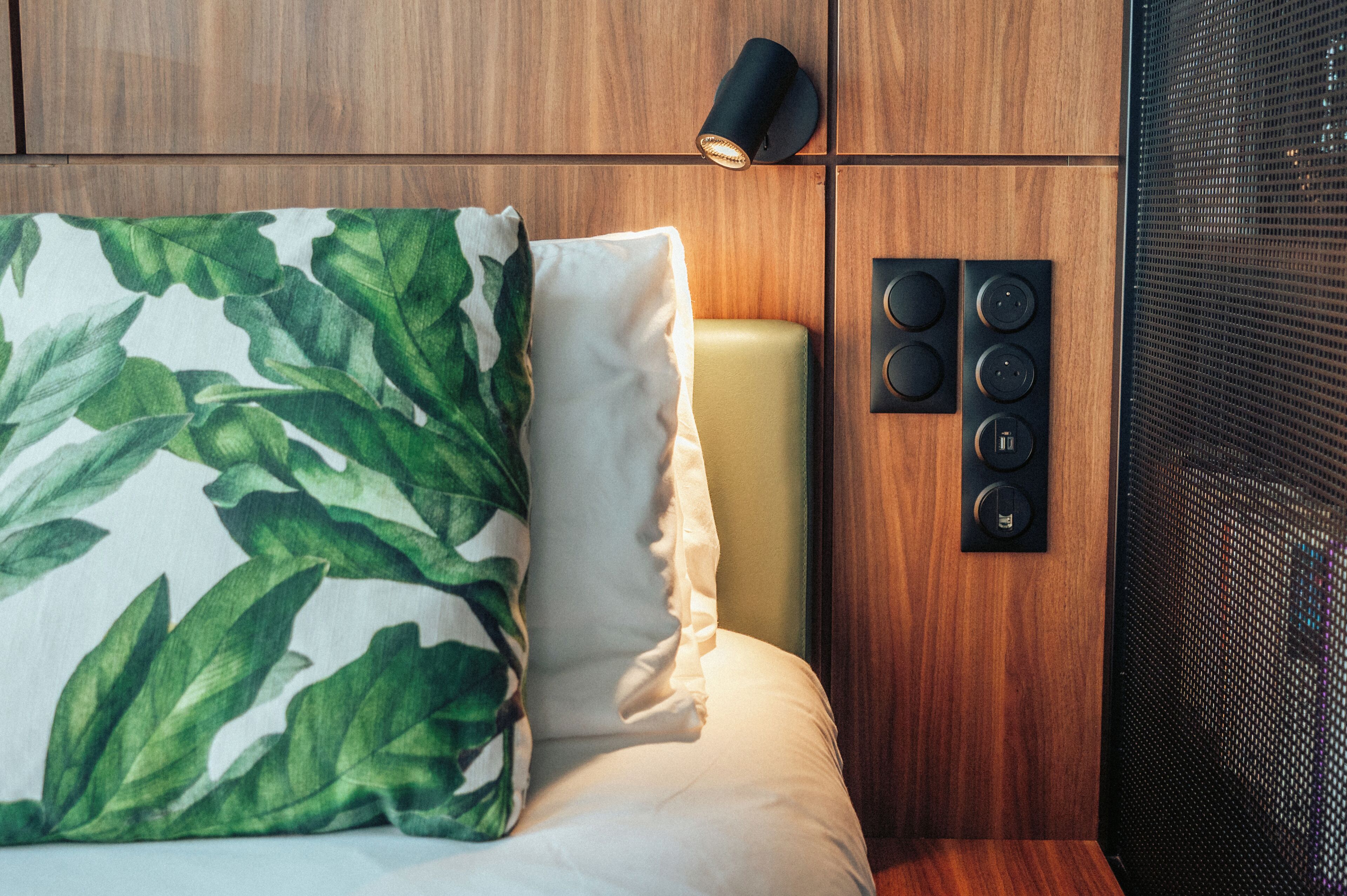 A hotel room with a bed, a green leaf-patterned pillow, a white pillow, a wall-mounted light, and a speaker system.