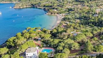 Vista aerea di una proprietà costiera con piscina, circondata da vegetazione lussureggiante e una spiaggia.