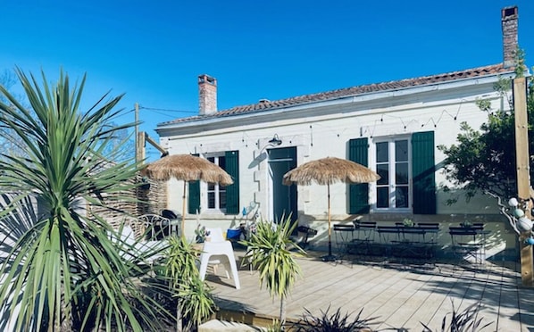 A white building with green shutters, a chimney, and a tiled roof. There are two thatched umbrellas, a white chair, and a table with chairs outside.