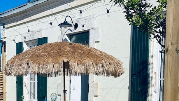Un patio al aire libre con una mesa y sillas, una sombrilla de paja y un edificio blanco con contraventanas verdes al fondo.