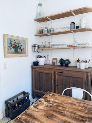 A rustic kitchen with wooden cabinets, a wooden table, and shelves holding kitchenware and plants.