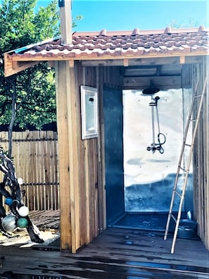 Struttura per doccia esterna in legno con tetto in tegole, una scala e un soffione sospeso.