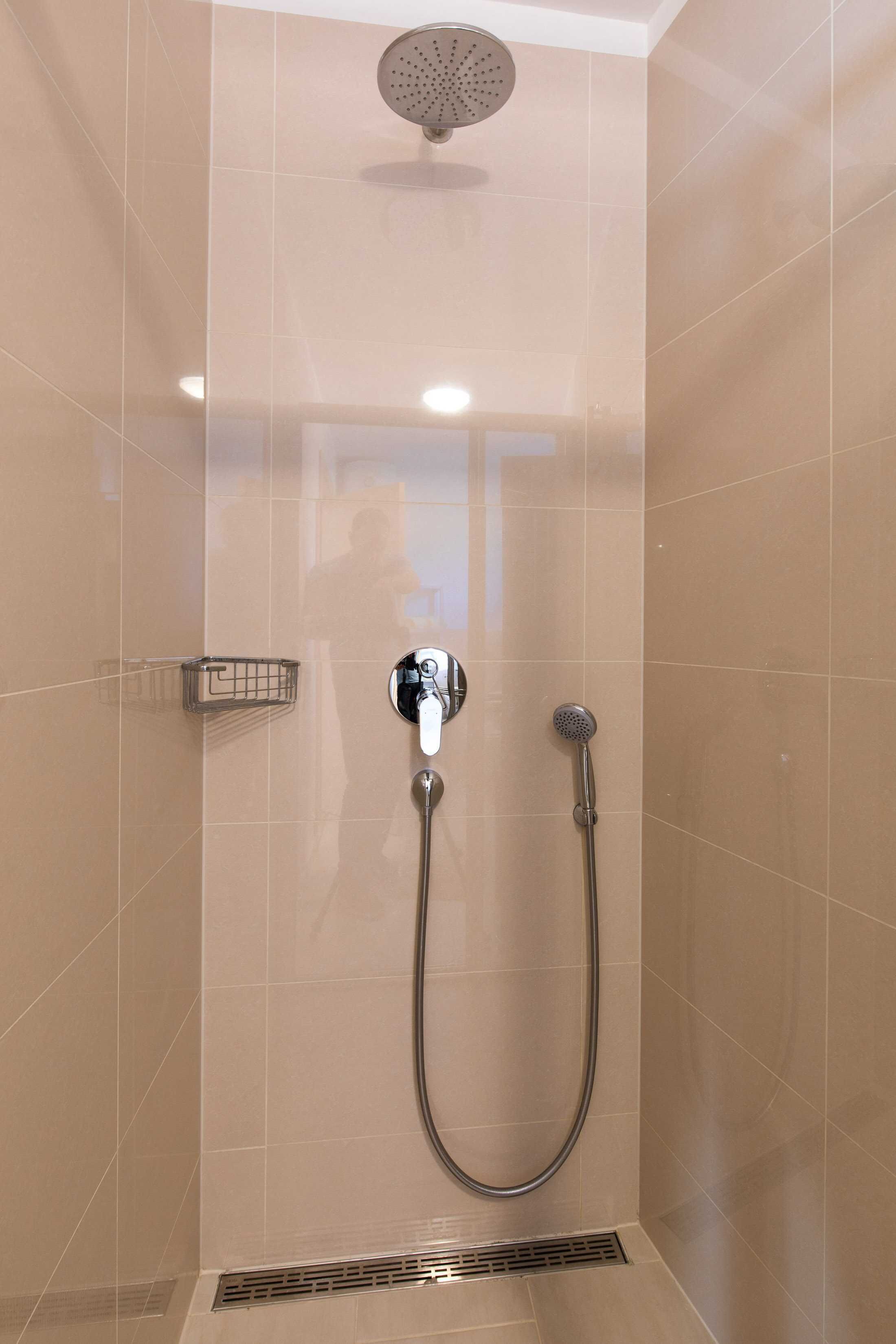 A shower with a handheld showerhead and a wall-mounted faucet, a small metal shelf, and a vent at the bottom.
