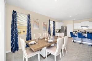 Un coin repas avec une table en bois dressée pour quatre personnes, des chaises blanches et une cuisine en arrière-plan.
