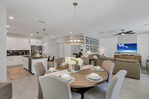 A modern kitchen with white cabinetry, a dining area with a wooden table and chairs, and a living area with a sofa and a television.